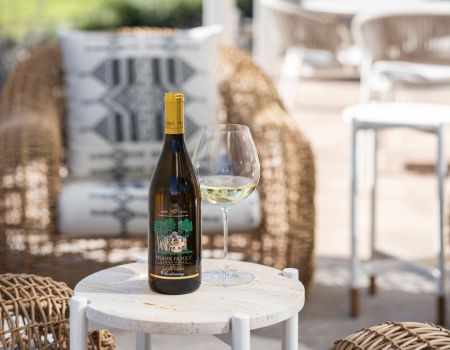 A bottle of Frank Family's Carneros Chardonnay next to a glass set in front of outdoor furniture