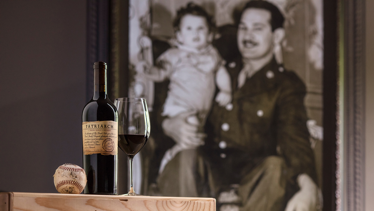 A baseball, wine bottle and glass of wine sit in front a picture of Hy Frank